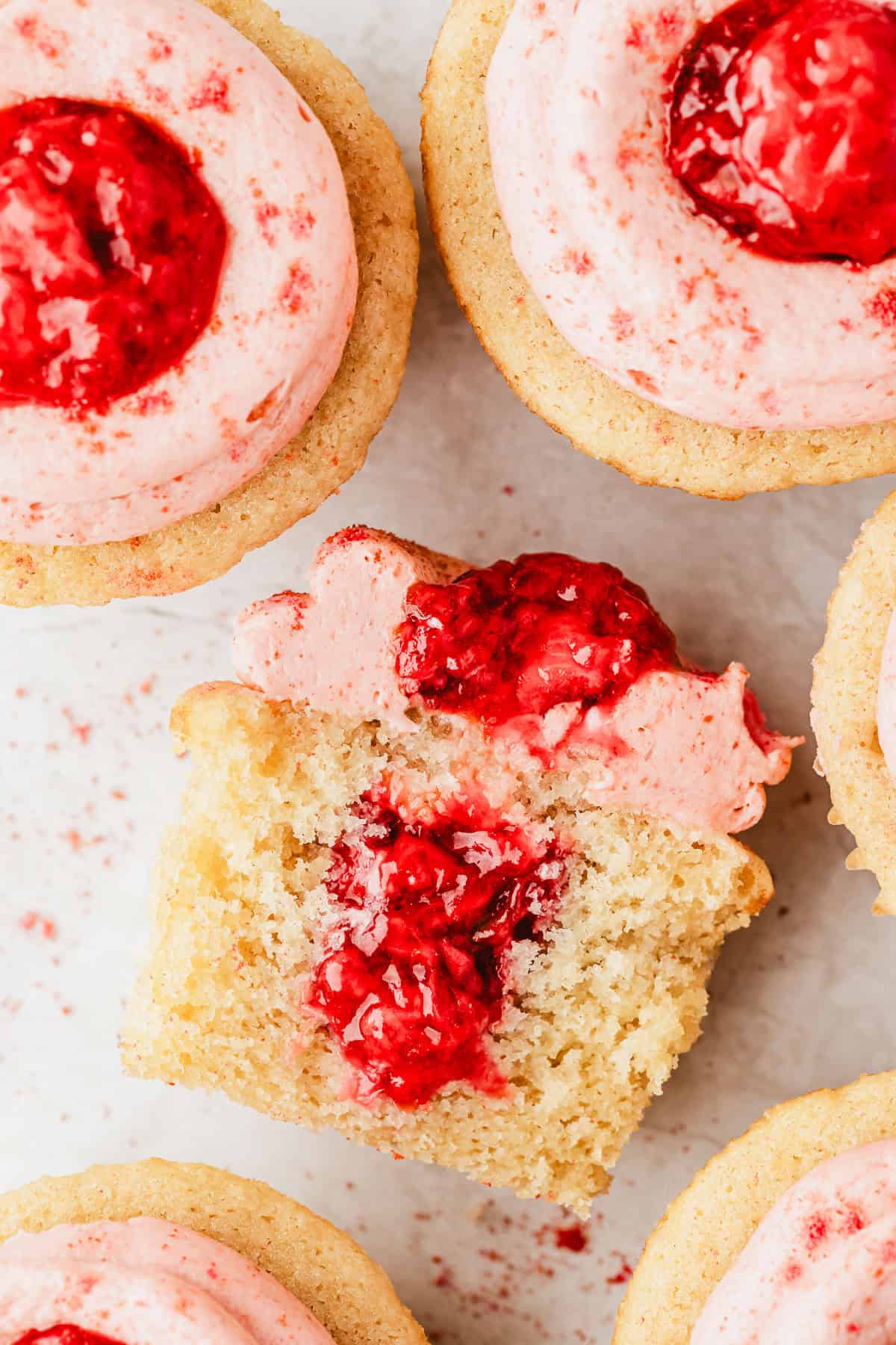 A cupcake with a bite taken out of it next to small batch strawberry filled cupcakes on a white counter.