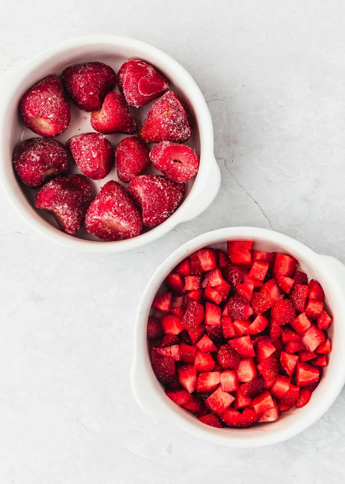 White bowls of frozen and fresh strawberries on a white counter.