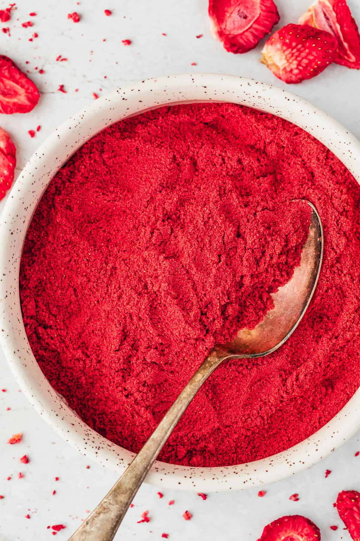 A white bowl of small batch homemade strawberry powder and a spoon on a white counter next to freeze dried strawberries.