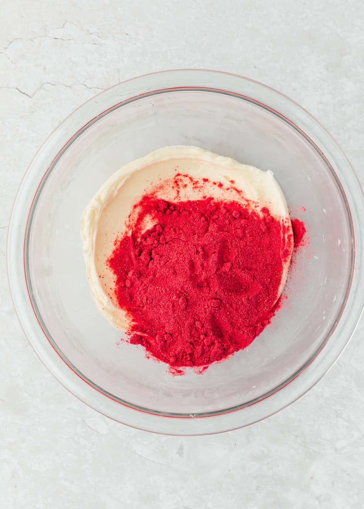 A glass bowl of frosting and small batch strawberry powder on a white counter.