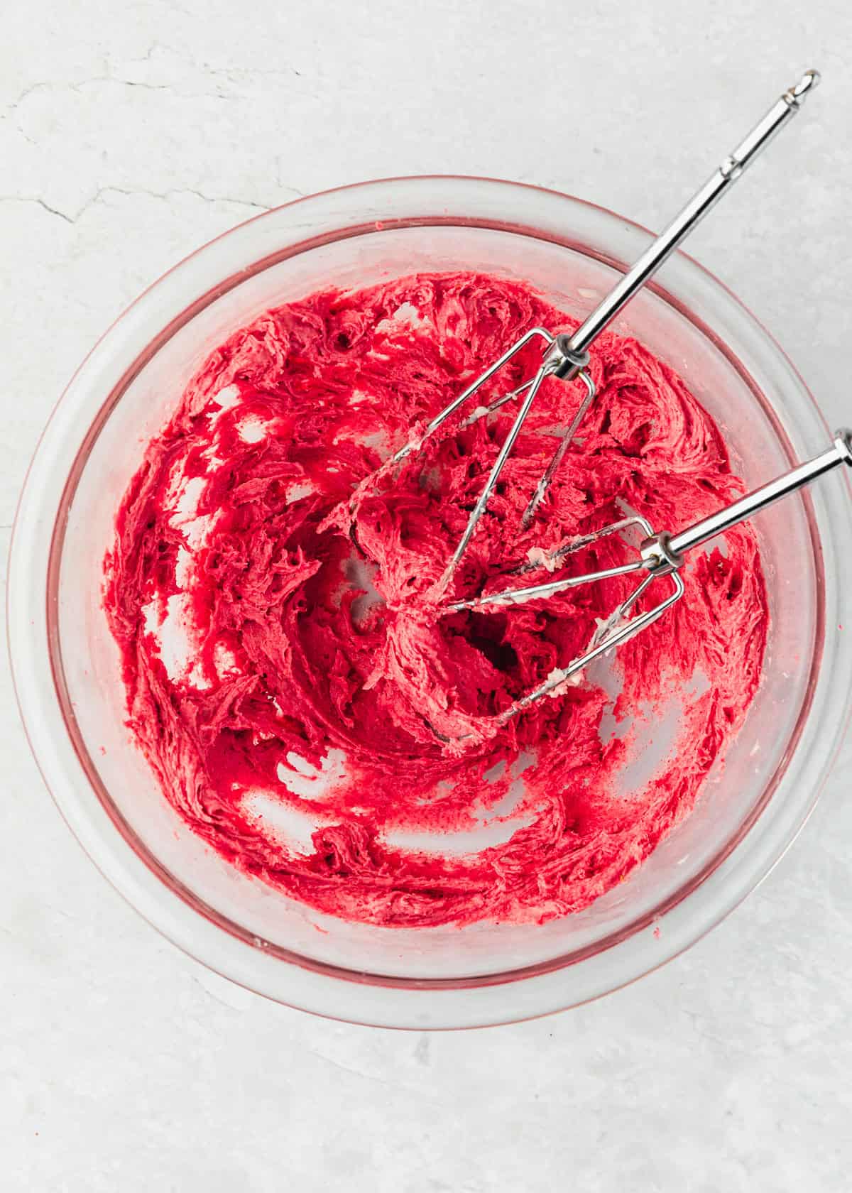 A clear bowl of strawberry frosting with beaters on a white counter.