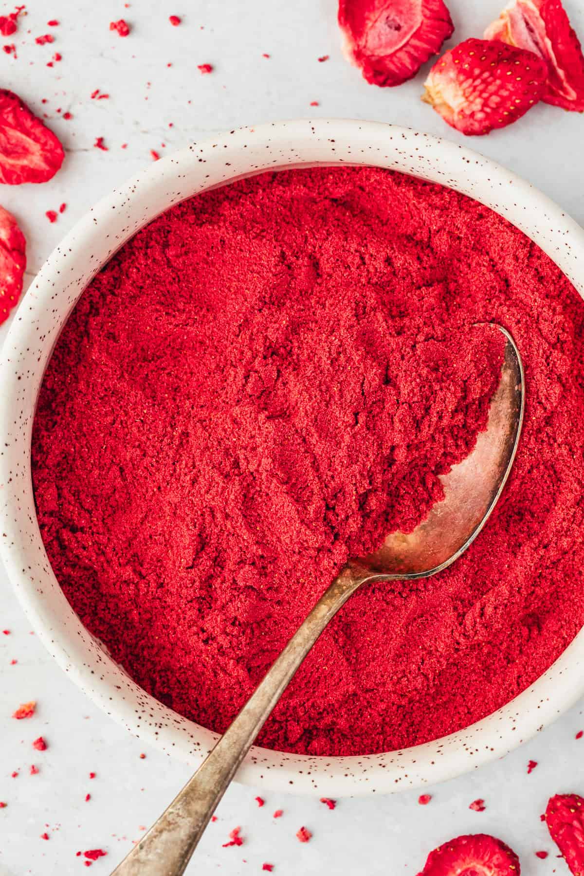 A white bowl of freeze dried strawberry powder next to freeze dried strawberries on a white counter.