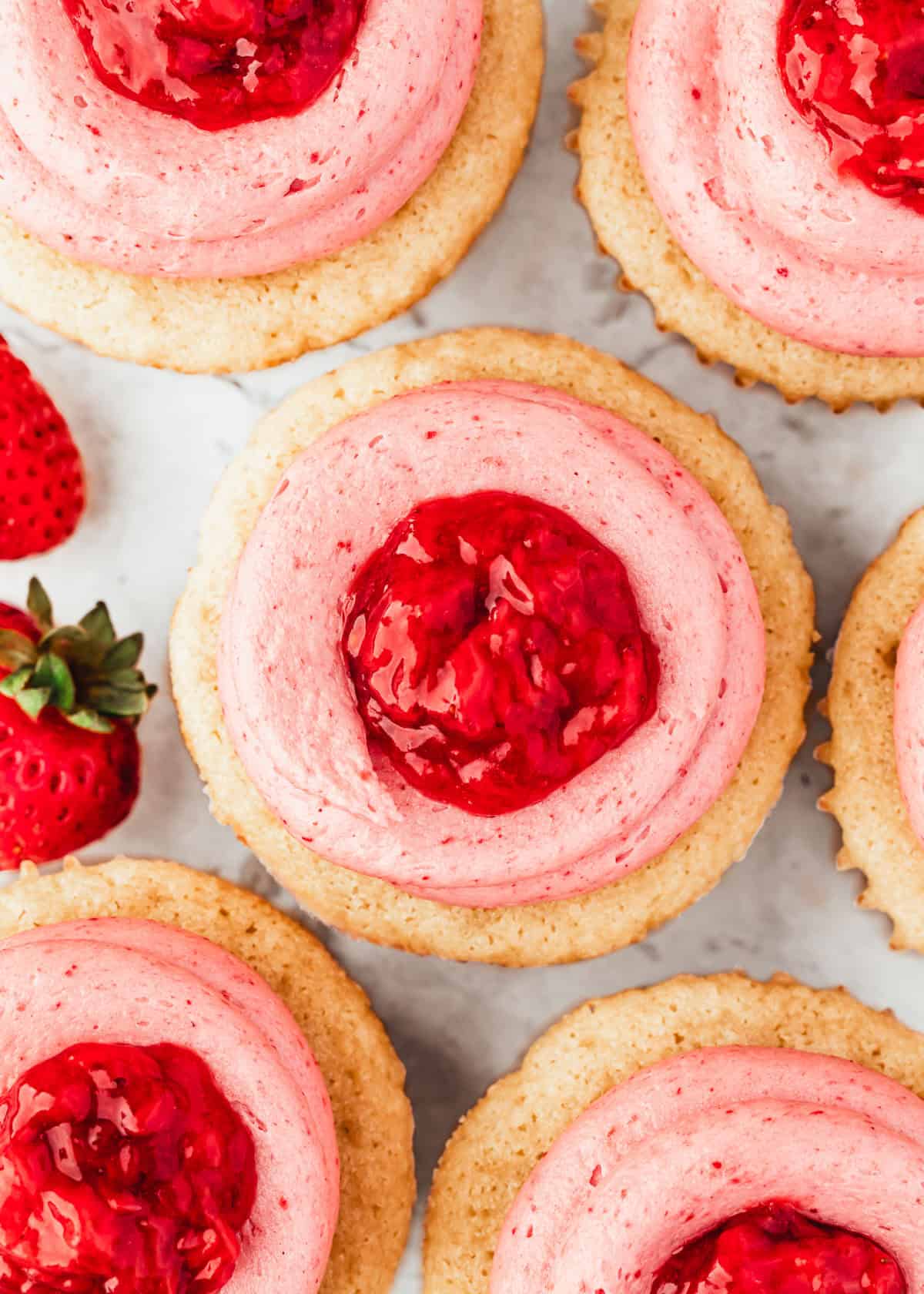 Rows of small batch cupcakes with strawberry buttercream next to strawberries on a white marble counter.
