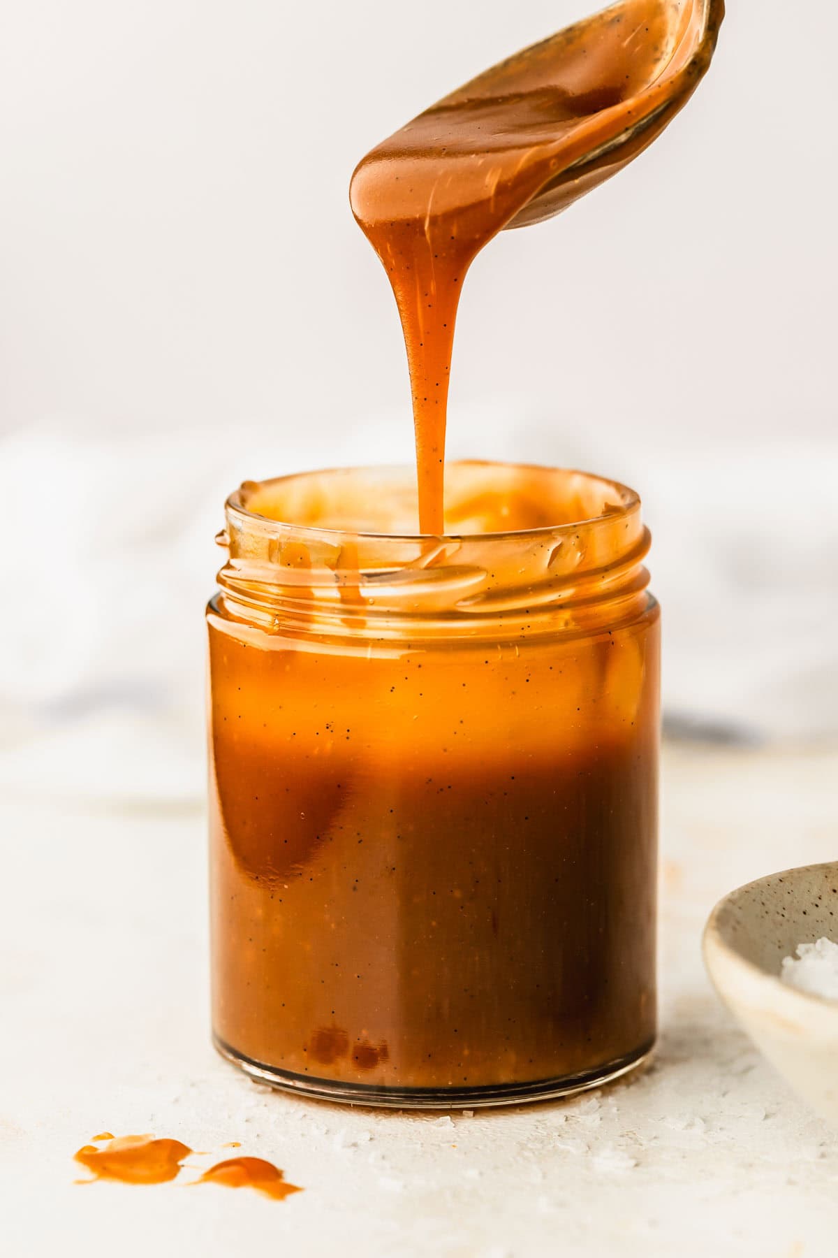 A spoon drizzling caramel into a jar of small batch caramel sauce on a white counter next to a white bowl of flaky salt.