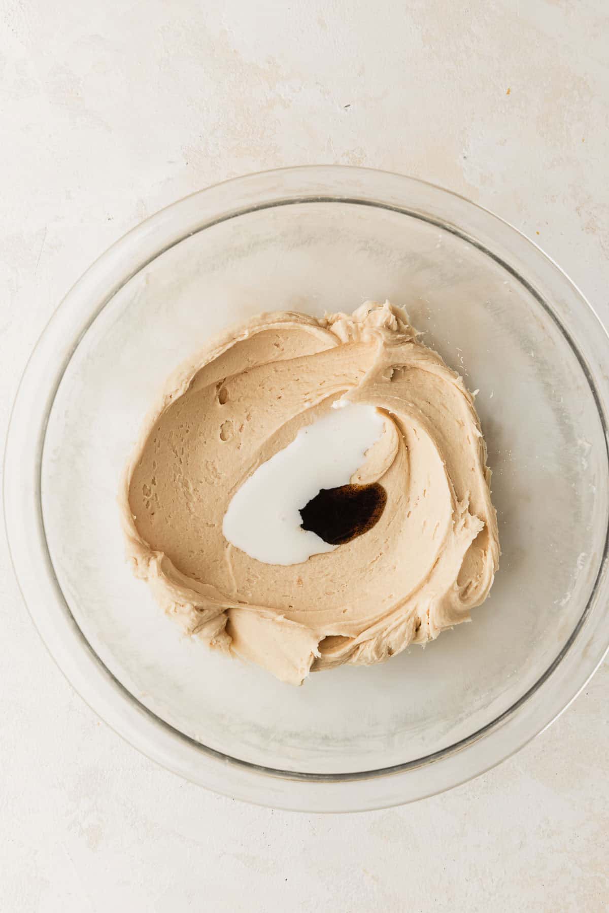 A clear bowl of small batch caramel buttercream frosting with milk and vanilla bean paste on a tan table.
