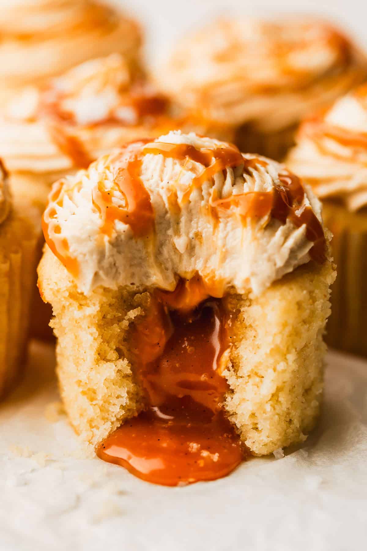 A cupcake with caramel dripping out next to rows of small batch caramel-filled cupcakes on parchment paper.
