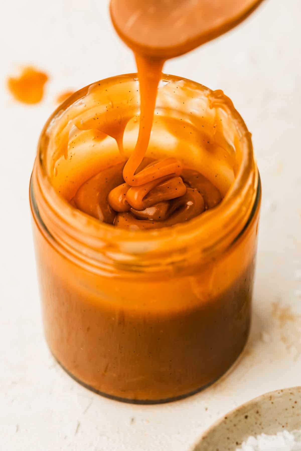A spoon drizzling small batch salted caramel sauce into a jar on a white counter.