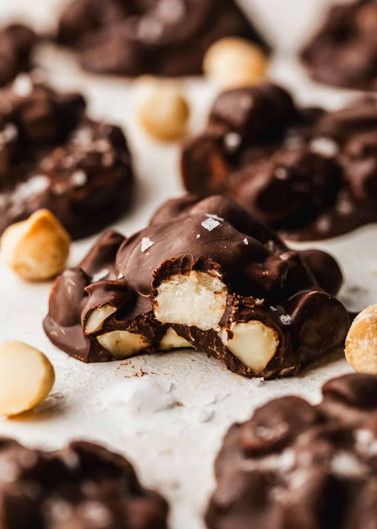 A chocolate with a bite taken out of it next to rows of small batch macadamia nut chocolates on a beige counter.