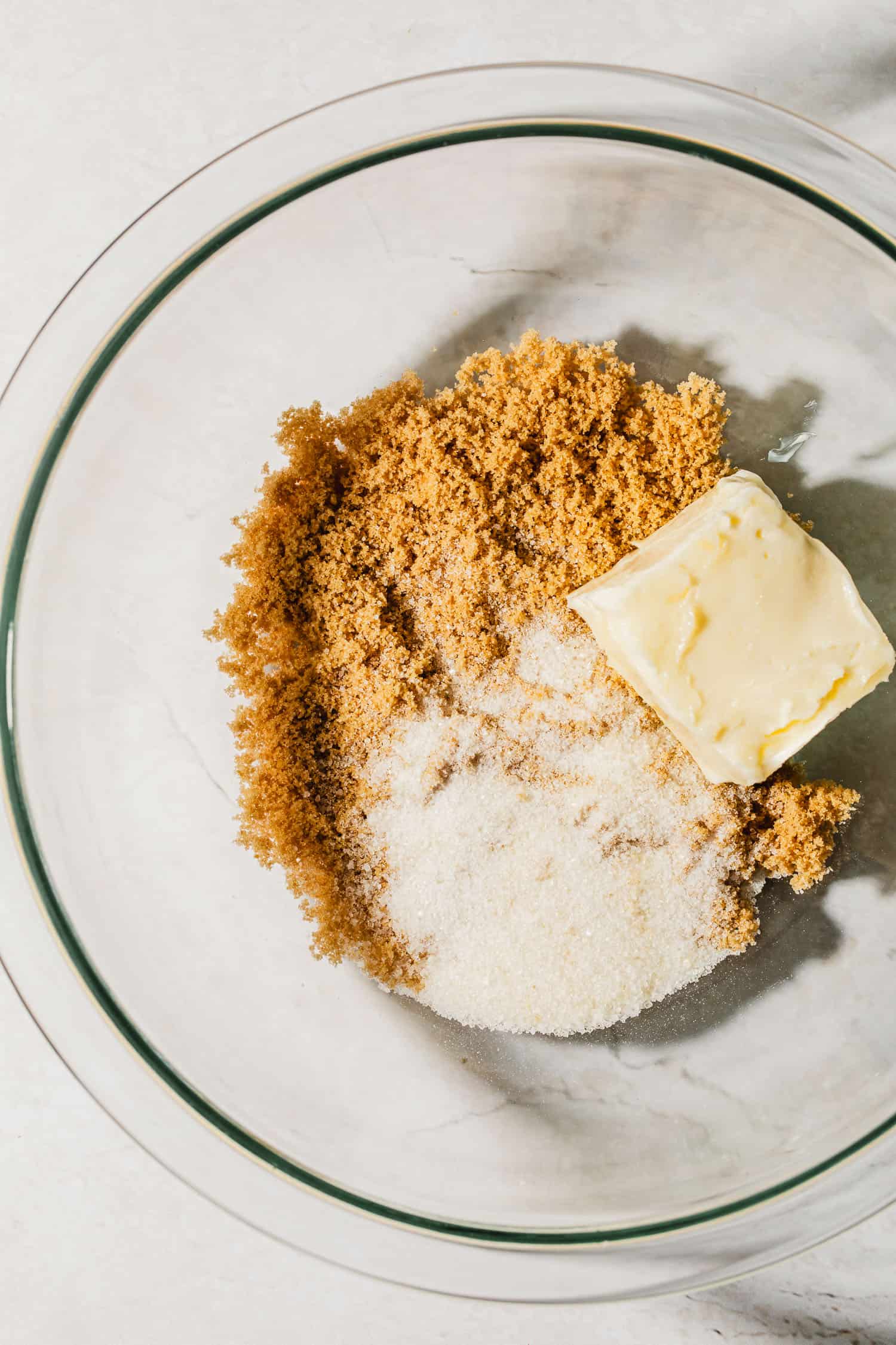 A glass bowl with brown sugar, granulated sugar, and butter on a white counter.