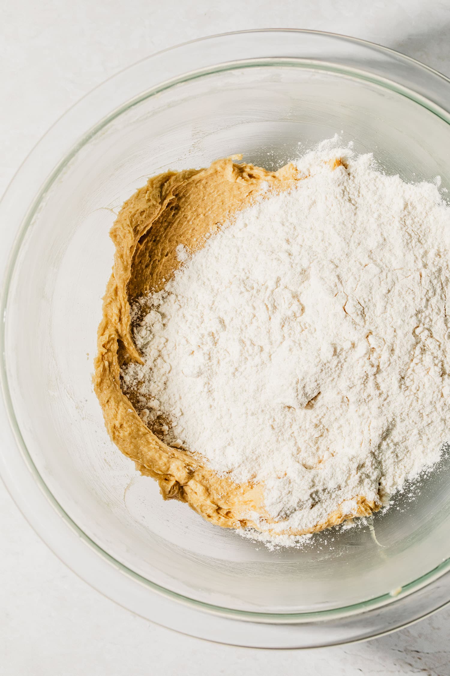 A glass bowl of creamed butter and sugar and flour on a white table.