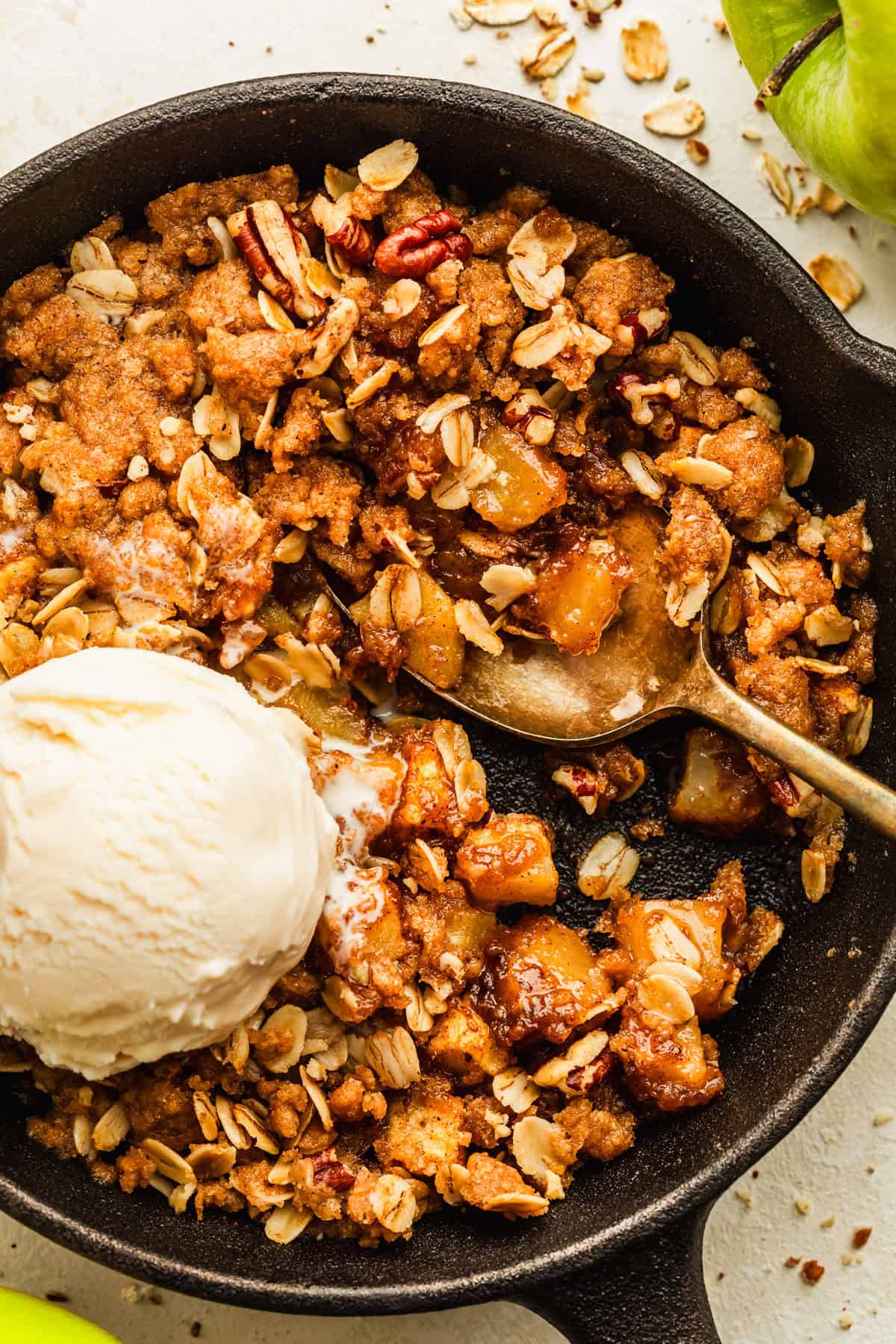 A cast iron skillet with a single serving of apple crisp and a scoop of ice cream next to green apples on a tan counter.