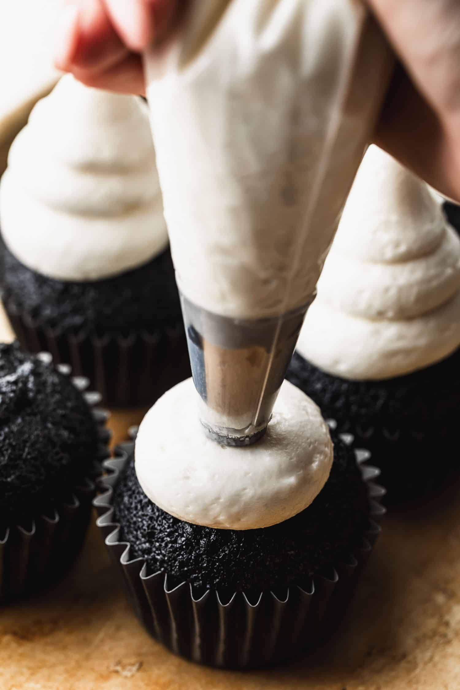 A hand using a piping bag to pipe white frosting on chocolate cupcakes.