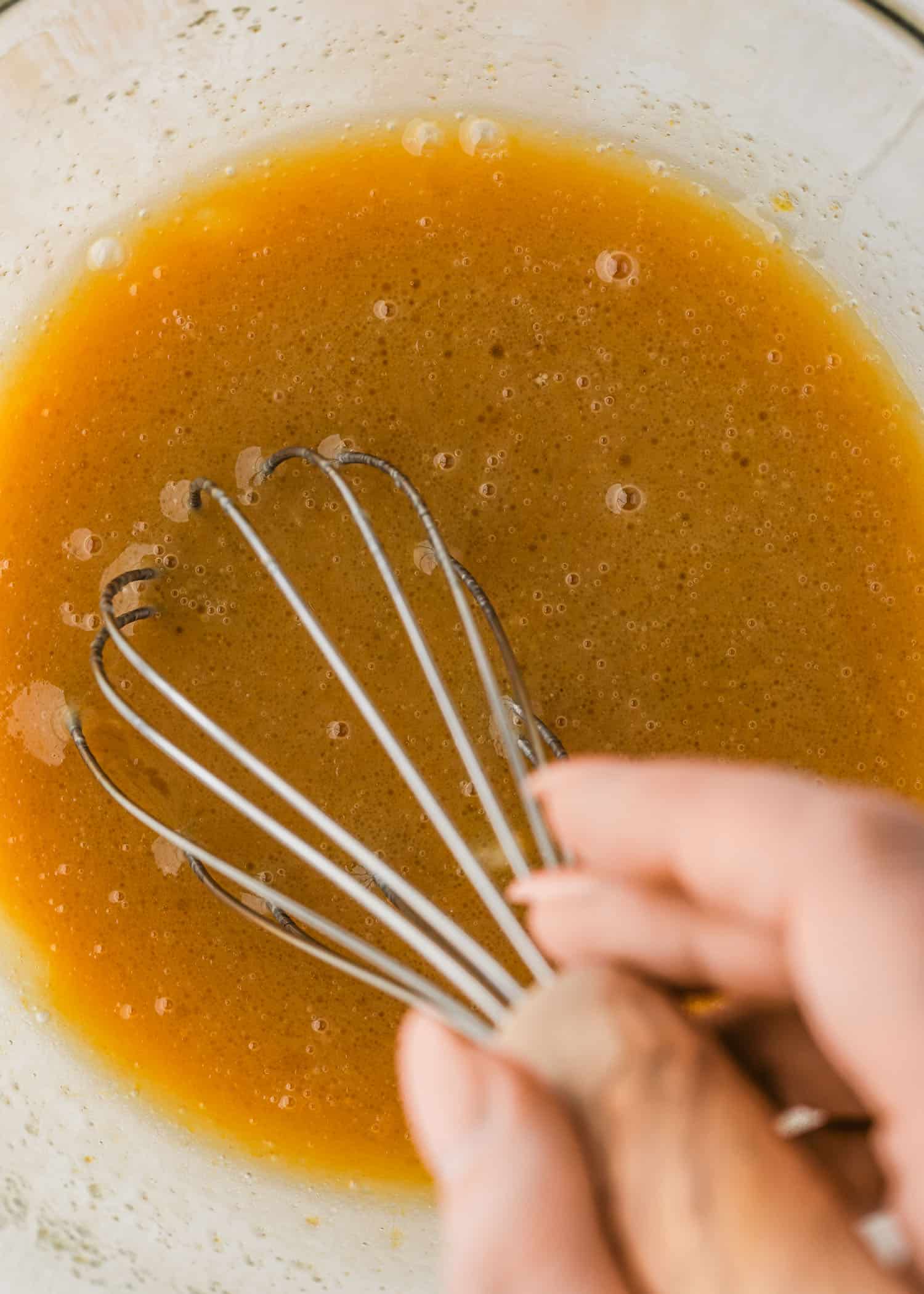 A hand using a whisk to mix wet ingredients in a clear bowl.