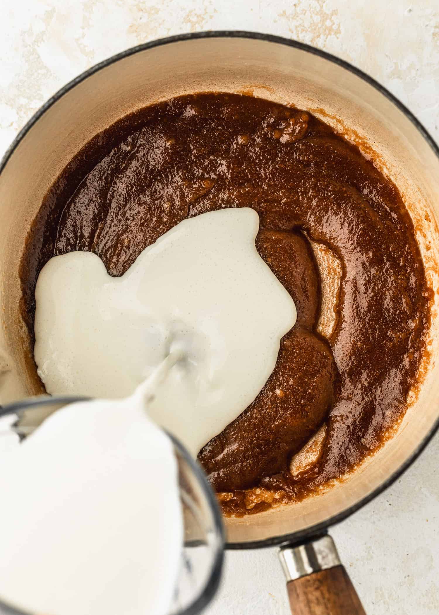 A hand pouring a cup of heavy cream into a white pot of brown sugar and butter.