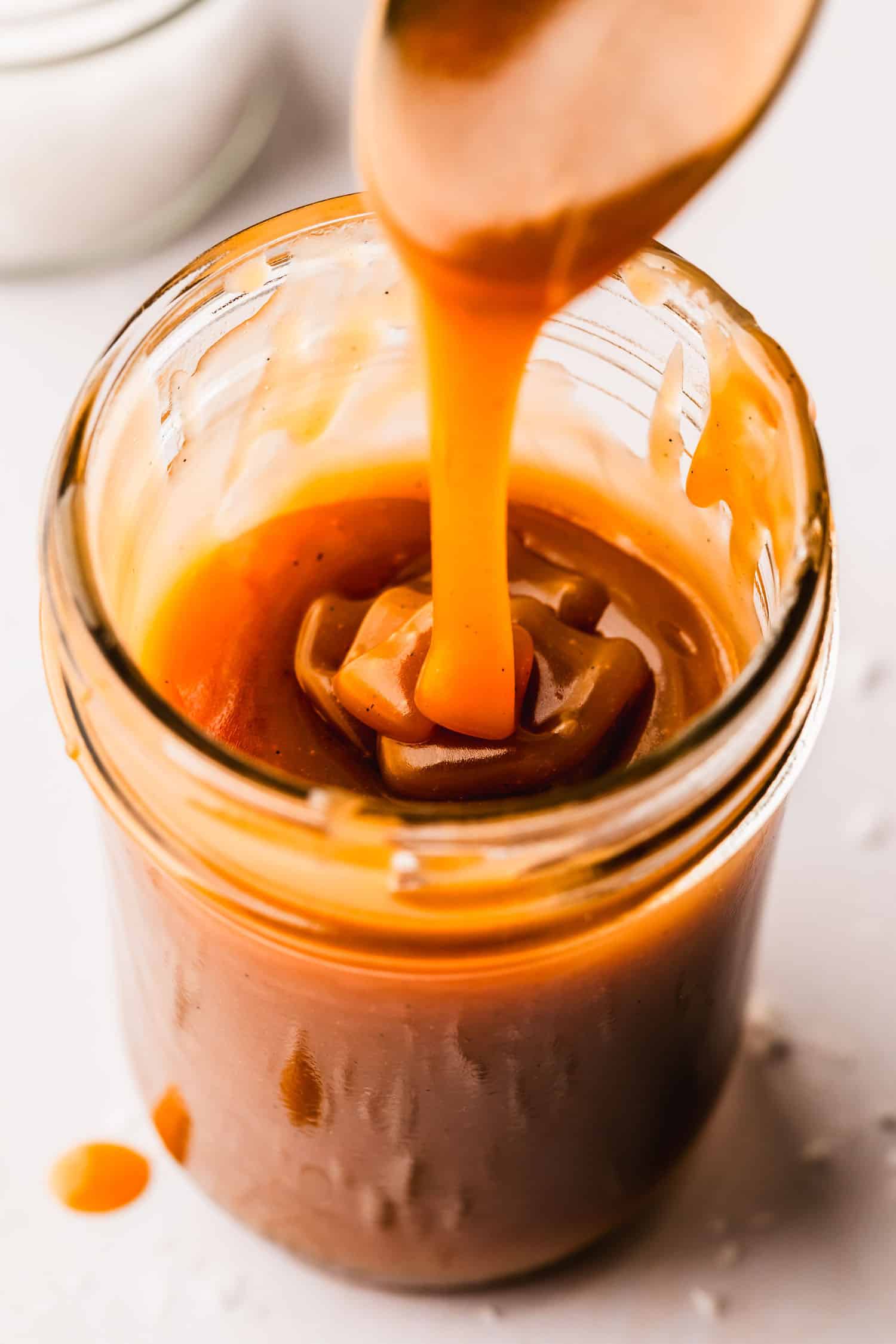 A spoon pouring small batch homemade butterscotch into a jar on a white counter.