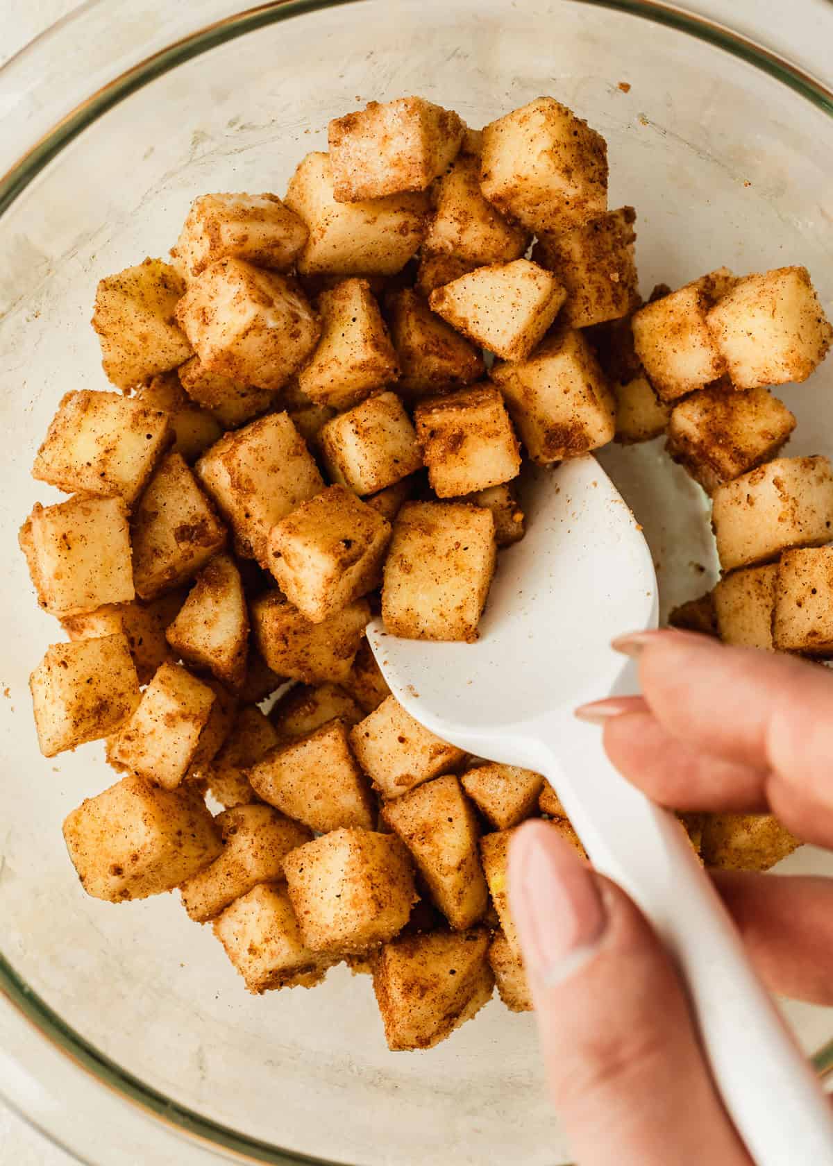 A hand using a rubber spatula to stir apples, spices, and brown sugar in a clear bowl.