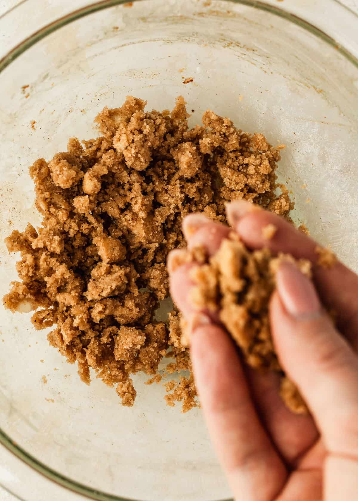 A hand crumbling butter, flour, and brown sugar in a clear bowl.